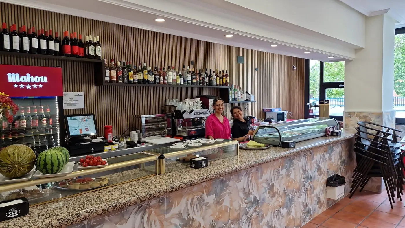 La barra del bar de lado a lado, con la cafetera, la vitrina de tapas, las botellas al fondo y dos camareras detrás