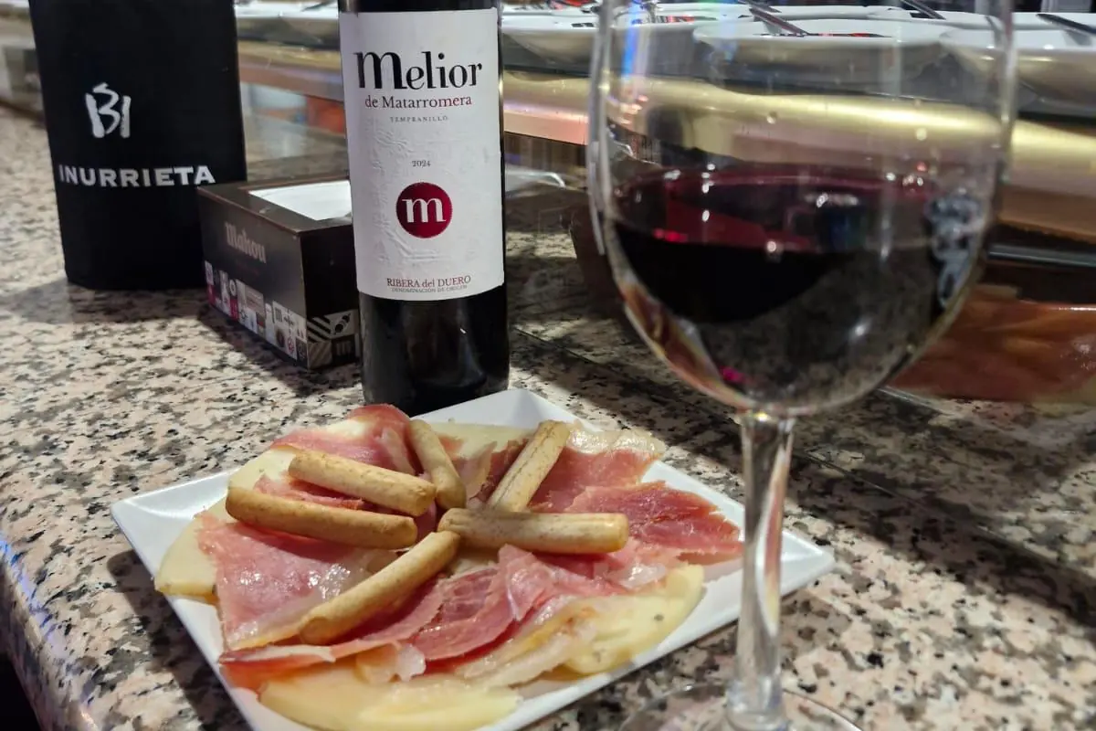 Copa de vino tinto y un plato de jamón y queso sobre la barra, con el cartel de Mahou al fondo